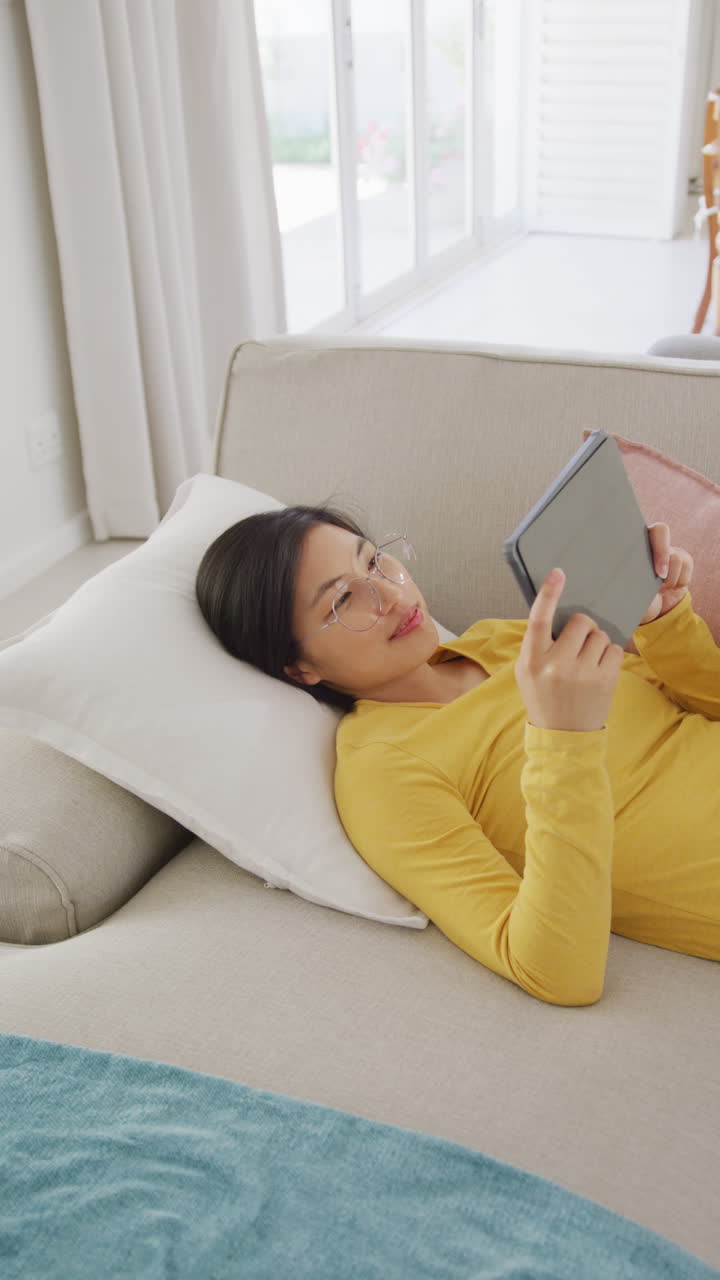 Vertical video of happy asian woman lying on coach and using tablet