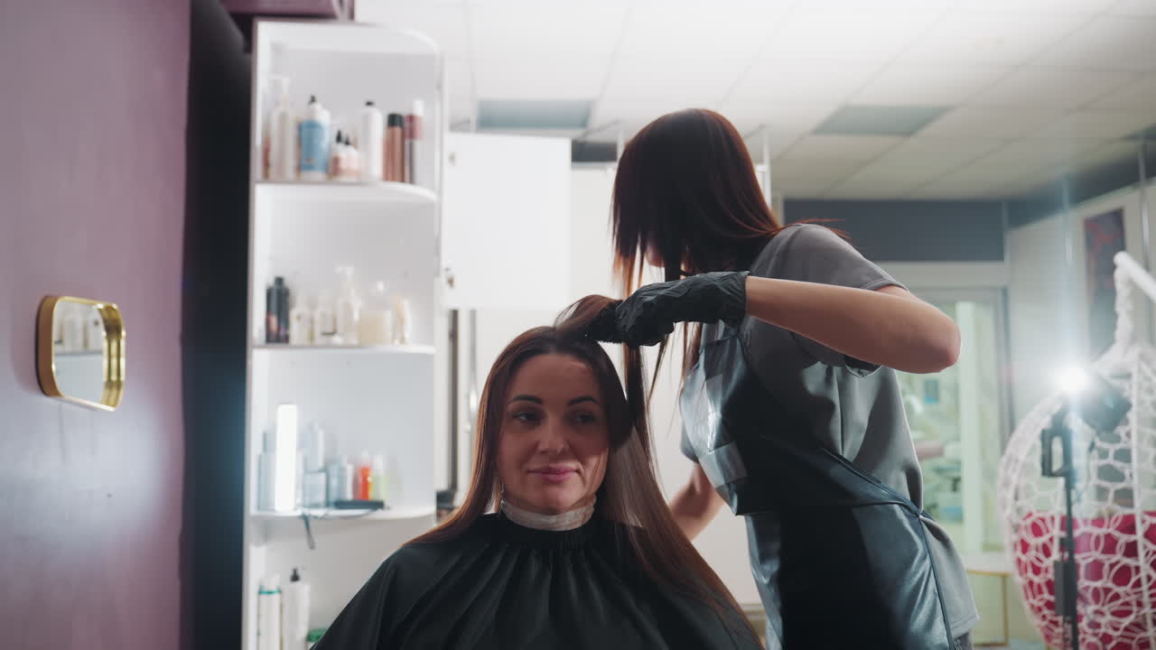 En colorist påfører farve på en kundes hår med pensel og handsker, omhyggelig sektionering og udglatning, skåle og fremkalder på nærliggende hylder, professionel kappe og fokuseret teknik under stærkt salonlys