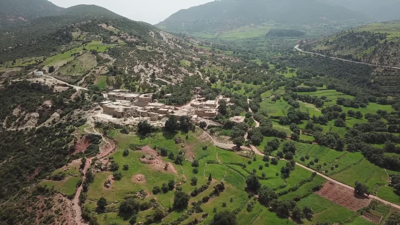 Ampia ripresa aerea panoramica di una lussureggiante valle verde sui monti dell'Atlante nordafricano. Il clip video cattura un villaggio rurale circondato da campi terrazzati e una tortuosa strada di montagna che collega l'area.