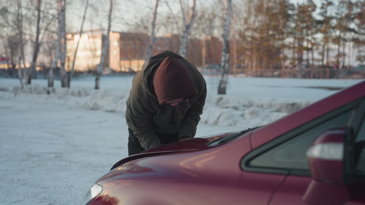 Student wearing hoodie bends over red car struggling to close bonnet on snowy ground with soft winter lighting, distant trees, and blurred residential buildings visible in background