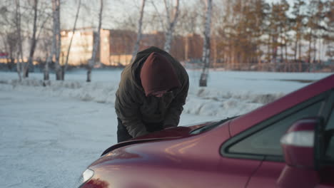 student wearing hoodie bends over red car struggling to close bonnet on snowy ground with soft winter lighting, distant trees, and blurred residential buildings visible in background
