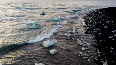aerial flyover black beach with small icebergs on shoreline during golden sunset, low altitude