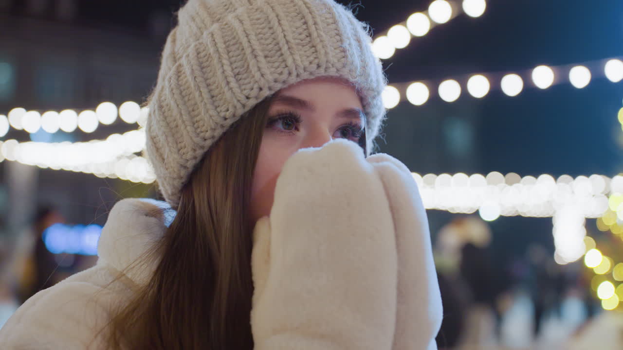 Young girl sits outdoors in winter, covering her face with gloved hands before exhaling visible cold breath, city lights glow behind her as people move around in a lively, festive urban environment