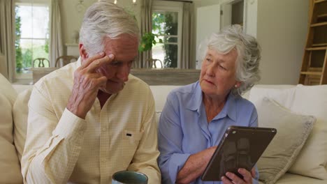 Estresada-Pareja-De-Ancianos-Caucásicos-Sonriendo-Mientras-Usa-Una-Tableta-Digital-Sentada-En-El-Sofá-De-Casa