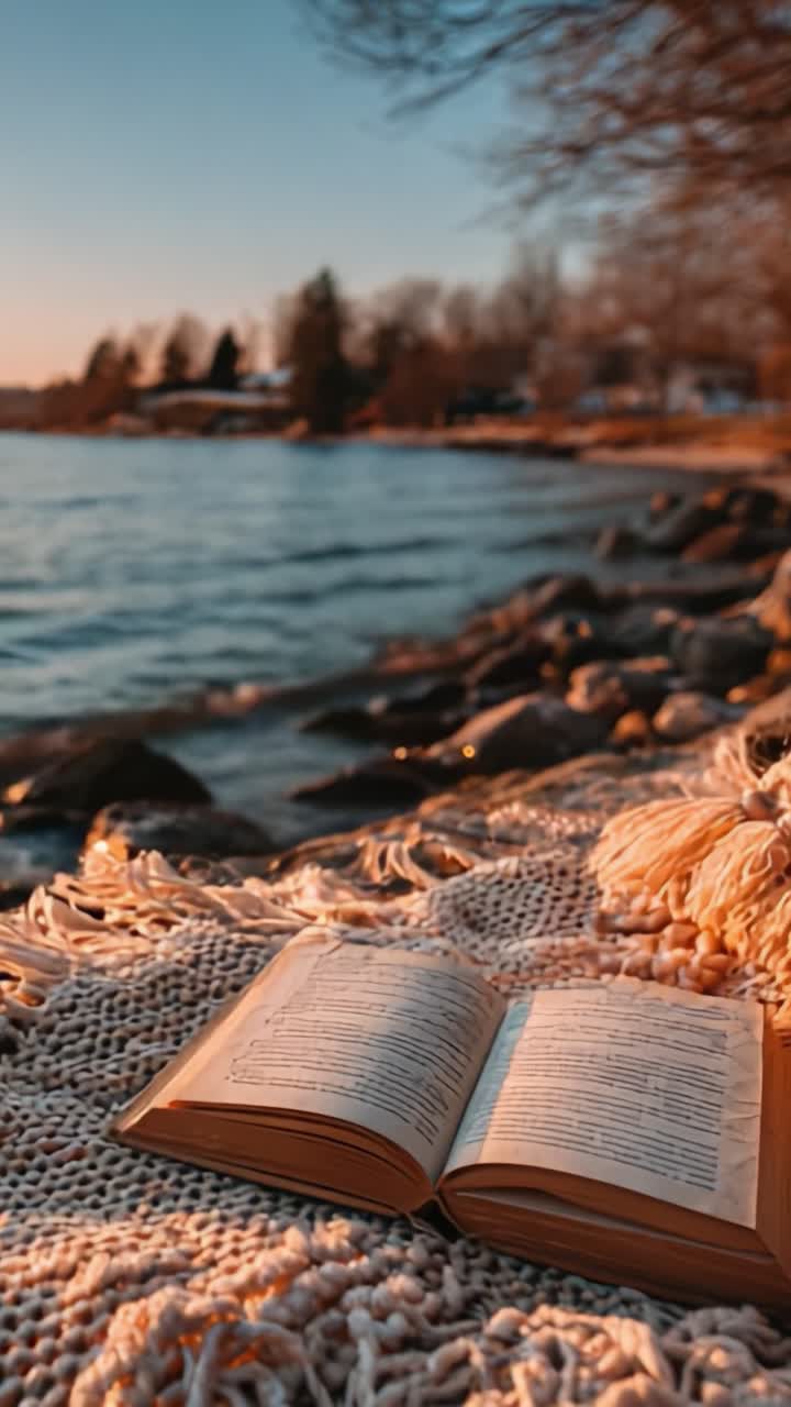 A tranquil moment by the water's edge, featuring an open book resting on a cozy textured blanket, surrounded by serene nature and sparkling waves at sunset