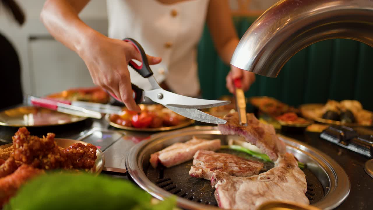 Person cuts grilled meat with scissors over tabletop grill, warm lighting, close-up, indoor dining