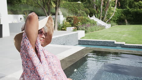 feliz anciana afroamericana en sombrero de sol sentada en el sol por la piscina, espacio de copia, cámara lenta