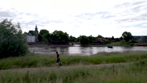 paso y seguimiento aéreo de un corredor de trail masculino en un camino fuera de carretera a lo largo del río ijssel en la llanura holandesa con el rostro de la ciudad de la torre zutphen detrás