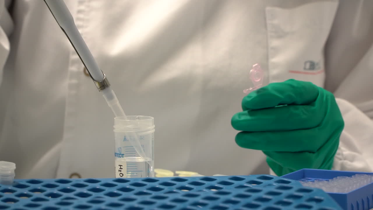 Scientist transfers liquid with pipette to tube held by green glove, close up