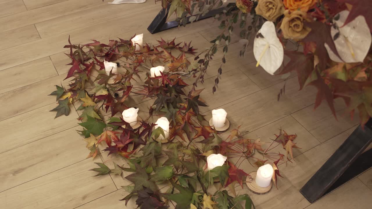 Guirnalda de hojas de otoño dispuesta en un suelo de madera con velas de pilar blancas y flores de temporada.