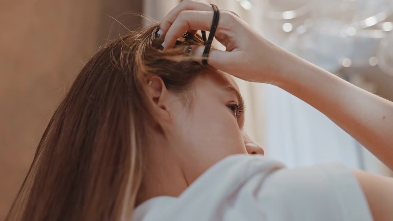 Focused woman fixing her hair near window. Woman concentrating on styling hair with gentle window illumination. Lady carefully arranging her hair while standing by softly lit window with focus
