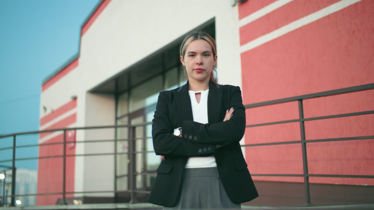 Young lady in black blazer confidently folds hands while standing in front of new modern building with red and white walls, expressing independence, and pride in personal achievement and growth