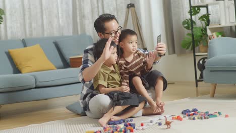 full body of asian father and sons playing the construction set colorful plastic toy brick at home. smiling and waving hello at smartphone, having video call, spending time together