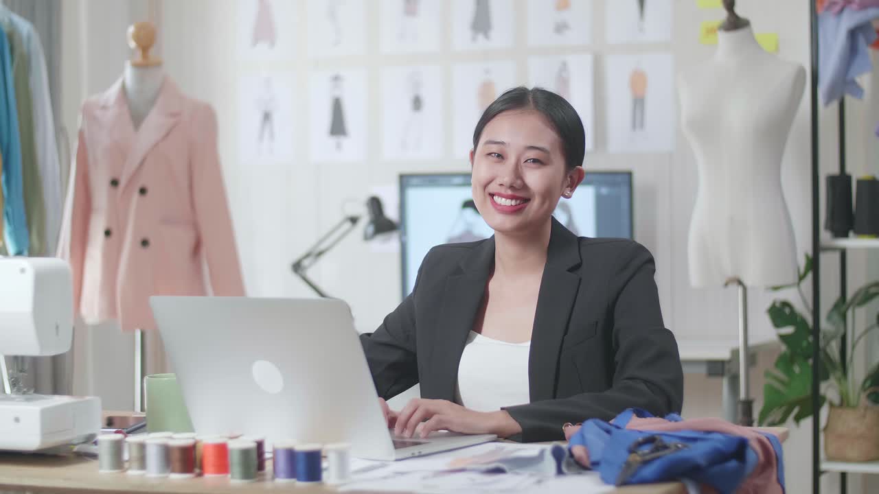 비즈니스 슈트를 입은 아시아 여성 디자이너가 노트북에서 일하고 스튜디오에서 옷을 디자인하는 동안 카메라에 미소 짓고 있습니다.