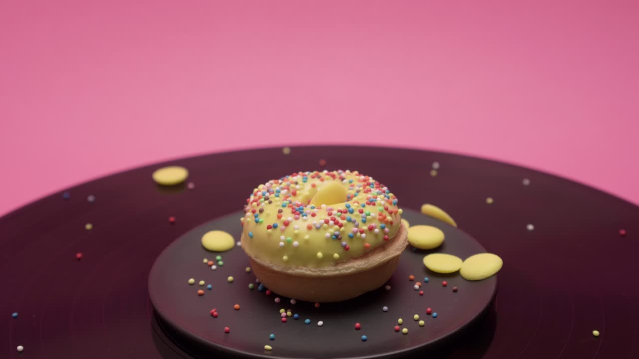 The American donut rolls in circles on a pink and black surface. It is topped with yellow chocolate and garnished with colorful crumbs and yellow choco disks