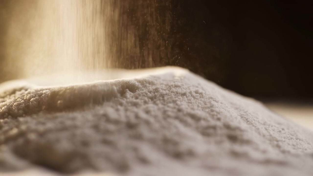 Pó branco fino assenta sobre uma pilha arredondada enquanto a poeira sobe através de uma luz bege quente, criando uma textura granular suave, fundo escuro e sombrio, perspectiva de close-up apertado e alimento minimalista e limpo.