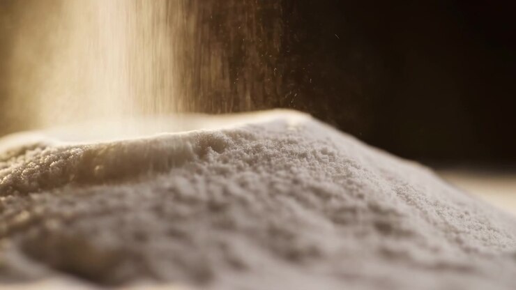 Fine polvere bianca si deposita su un cumulo arrotondato mentre la polvere si solleva attraverso una luce beige calda, creando una texture granulare morbida, sfondo scuro e ombreggiato, prospettiva ravvicinata e stretta, ed estetica alimentare pulita e minimale