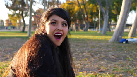 una linda chica sentada en un parque con hojas de otoño mirando hacia arriba con una mirada sorprendida y luego riéndose a cámara lenta