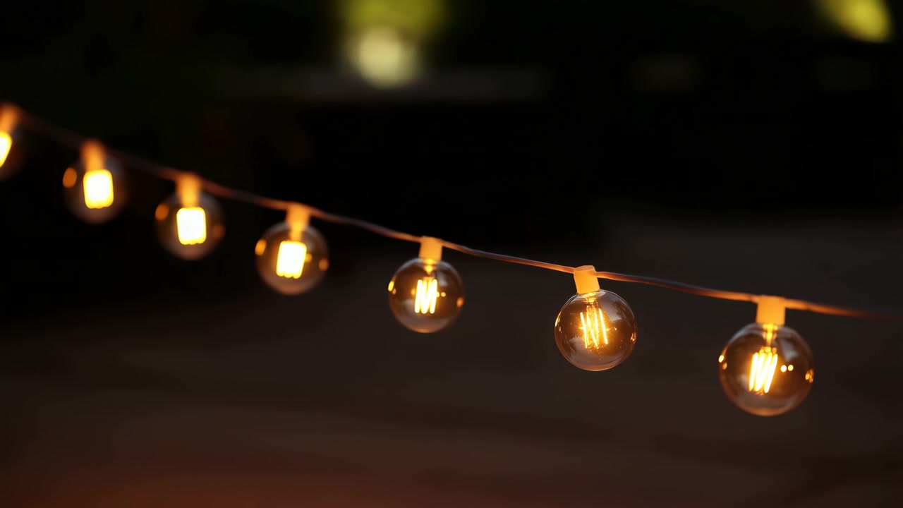 Pemandangan bergerak jarak dekat bola lampu bulat bening yang digantung pada kawat tipis gelap, bersinar dengan pantulan filamen hangat dengan latar belakang senja yang lembut dalam pengaturan dekorasi luar ruangan yang mengundang.
