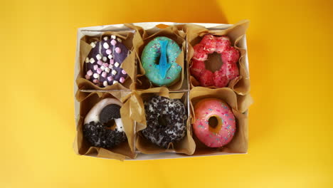 overhead shot of a hand picking a vibrant donut from a box filled with six assorted colorful donuts, each with unique toppings, against a bright yellow background.