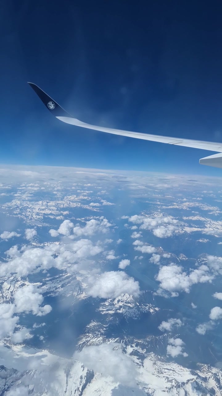 Utsikt fra et flyvindu viser vingen over en snødekket fjellkjede delvis skjult av spredte skyer, vertikal