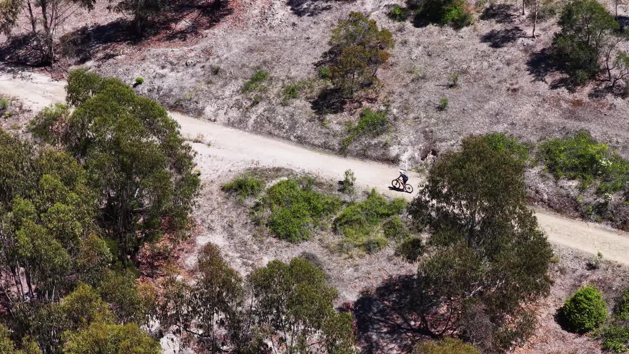 Uma tomada de drone em ângulo alto acompanha um ciclista solitário pedalando por uma paisagem australiana seca e arborizada