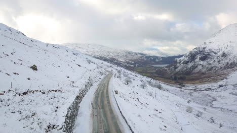 Aerial-footage-of-a-snow-covered-road-in-Snowdonia-looking-towards-Llyn-Gwynant-and-Nant-Gwynant,-Wales