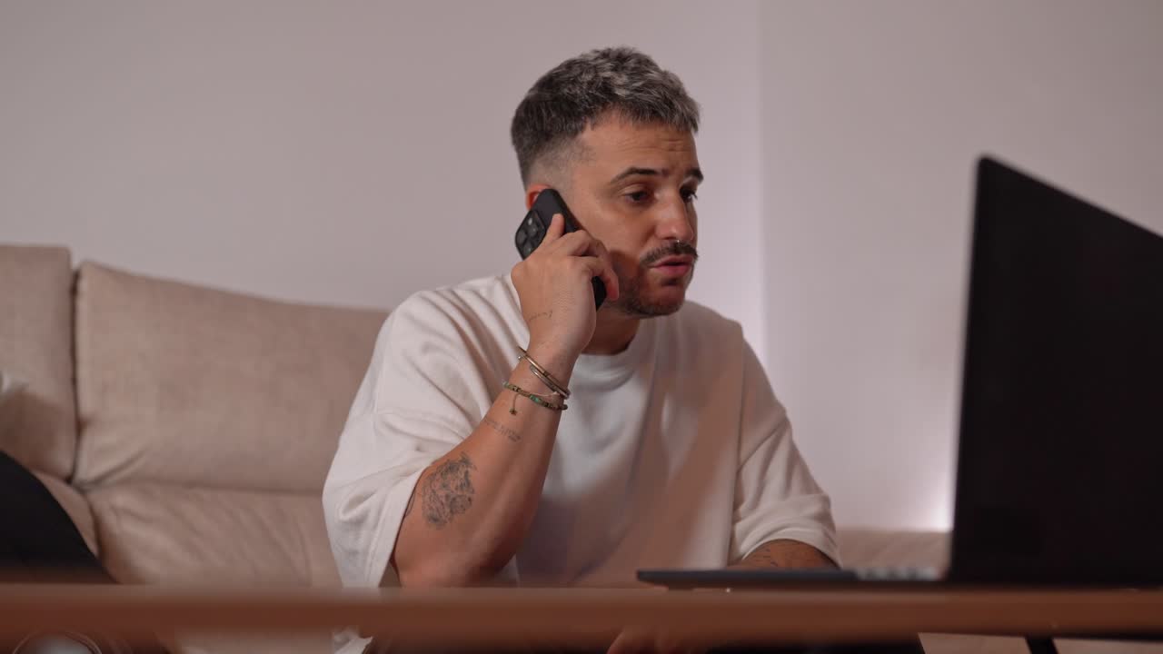 Man sitting on sofa having serious phone conversation while gesturing with hand in front of laptop at home