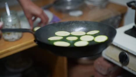 cut pieces of green zucchini placed by hand in black fry pan with hot oil, filmed as closeup slow motion shot