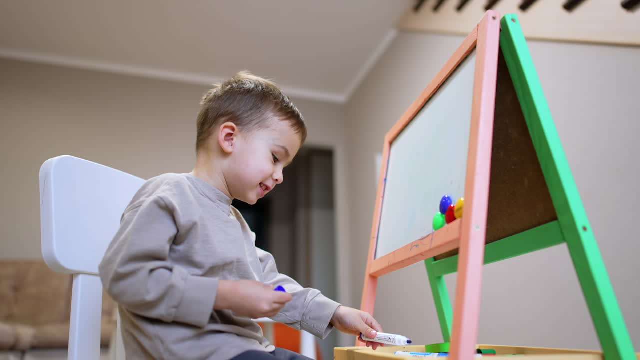 Bambino che disegna all'interno. Un bambino piccolo è seduto davanti a un cavalletto colorato, pensieroso e pronto a disegnare in una stanza accogliente e ben illuminata.