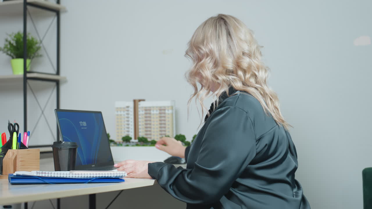 Side view of focused woman in black outfit seated at desk, opening laptop and reaching for mouse while surrounded by office supplies, coffee, notebooks, and building model