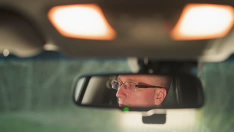 el rostro de un hombre se refleja en el espejo retrovisor de un automóvil, mostrando su expresión enfocada mientras usa gafas. la suave iluminación interior del vehículo destaca su comportamiento reflexivo