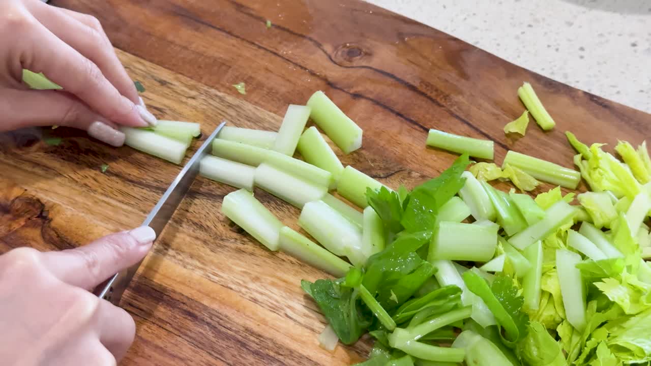 Hands use a kitchen knife to cut celery stalks into pieces on a wooden cutting board in a brightly lit kitchen, overhead perspective