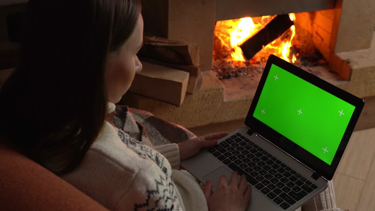 Woman at home, sitting on a sofa near the fireplace, working on a laptop computer with a green screen layout