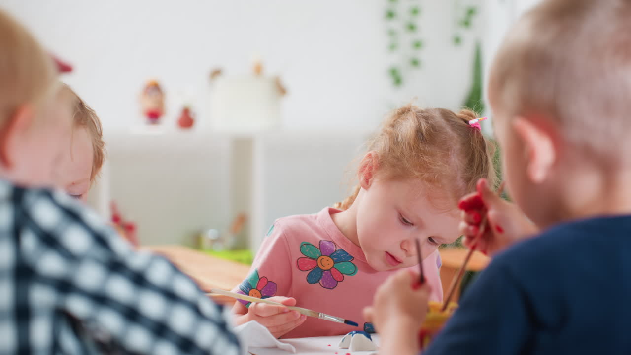 Close up rear view of boy in blue pulling paint container toward himself during art class as girl in pink shirt with flower design attentively watches, children concentrating on creative activity
