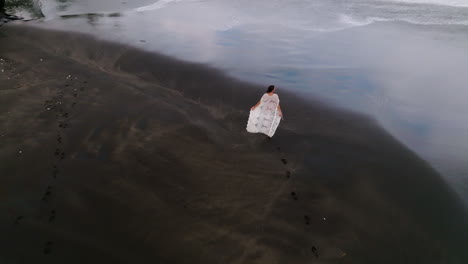 aerial drone shot of young woman walking on black sand beach in bali, indonesia