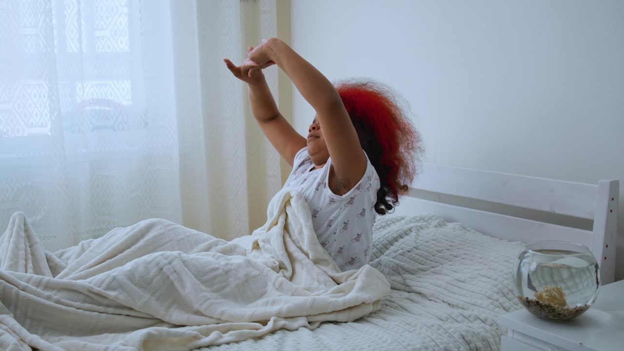 Relaxed african american preteen girl with vibrant red curly hair stretching lazily in bright white bedroom, enjoying peaceful morning sunlight and sense of well being