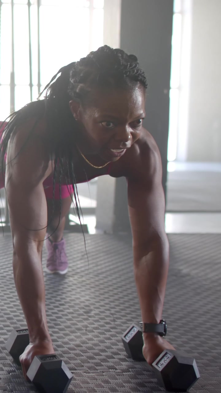 Vídeo vertical: Mulher afro-americana fazendo remada com halteres hexagonais em posição de prancha no colchonete na academia para fitness