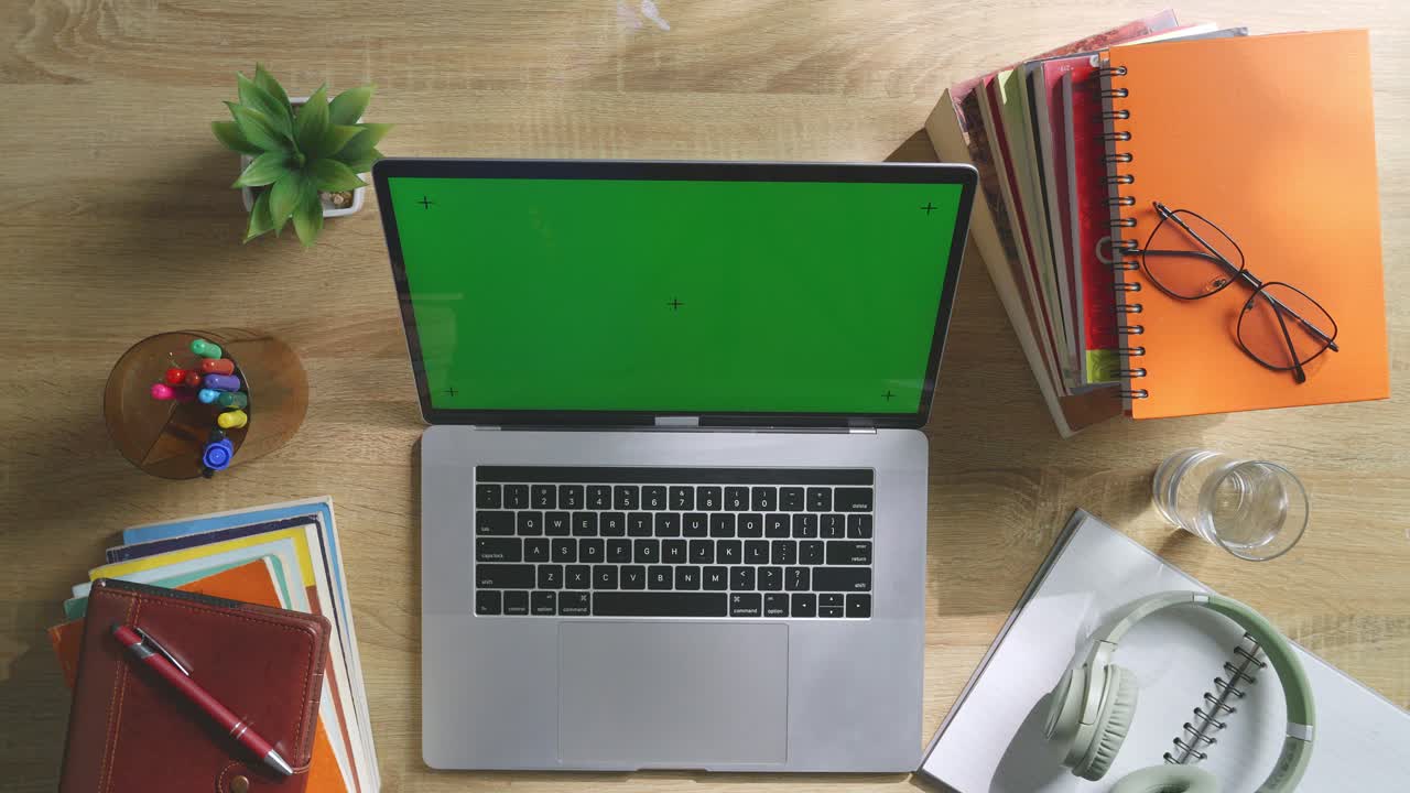 vista de arriba hacia abajo de una computadora portátil con pantalla verde simulada pantalla chromakey en un escritorio de oficina de madera al lado de la libreta con bolígrafos, gafas y un vaso de agua.