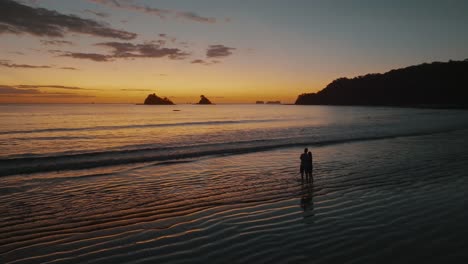 Silhouette-Of-Couple-Standing-At-The-Beach-During-Sunset-In-Guanacaste,-Costa-Rica---aerial-drone-shot