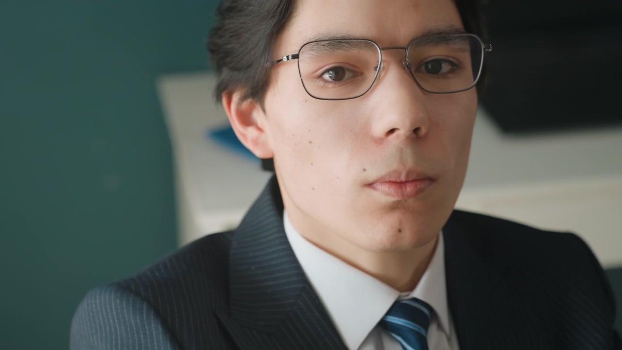 デスクで眼鏡を調整し、ノートとラップトップを使ってプレゼンの練習をする若いアジア人ビジネスパーソン。紺色のスーツとストライプのネクタイを着用し、真剣に集中しながらも微かな不安を抱え、書類を整理し準備を進めている。