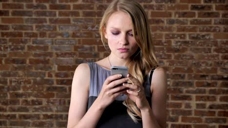 joven chica rubia linda está viendo fotos en el teléfono inteligente, concepto de comunicación, fondo de ladrillo
