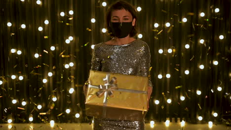 mujer joven con mascarilla y vestido de noche sosteniendo una caja de regalo dorada y confeti de brillo cayendo