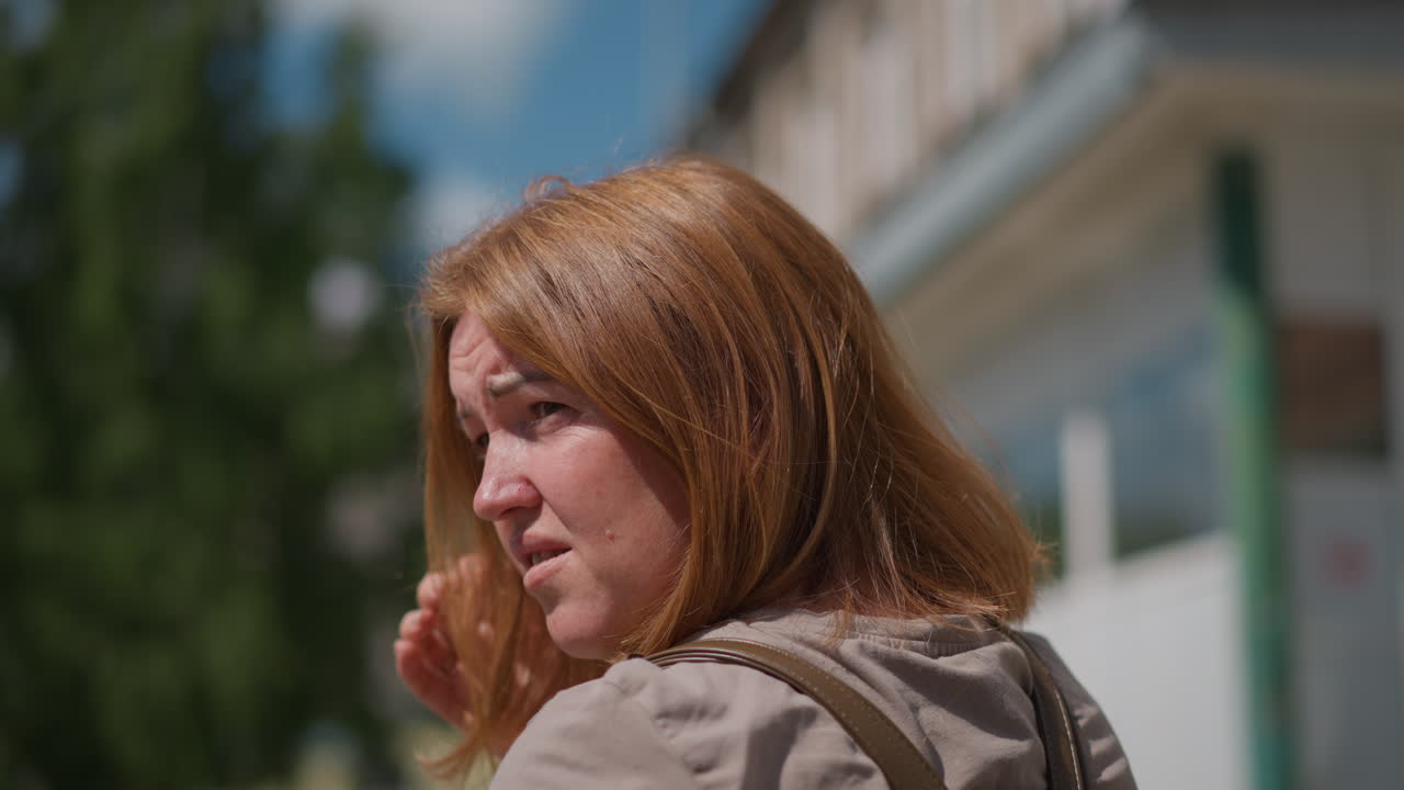 Tense woman seen in profile adjusting hair with hand wearing smart watch, standing near residential building in bright sunlight, subtle gesture expresses unease, and distracted emotional state