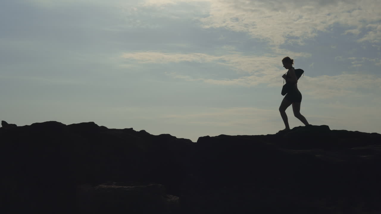 Silhouette of a woman walking along a rocky coastline at sunset