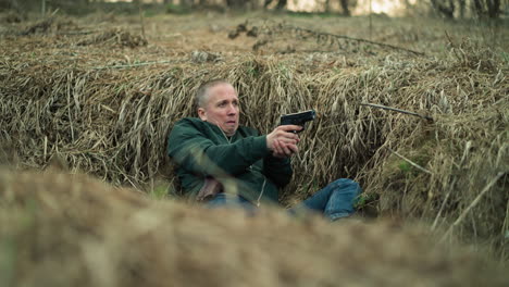 a close view of a man wearing green jacket and blue jeans lying in dry grass while holding a handgun and aiming with it, looking tense