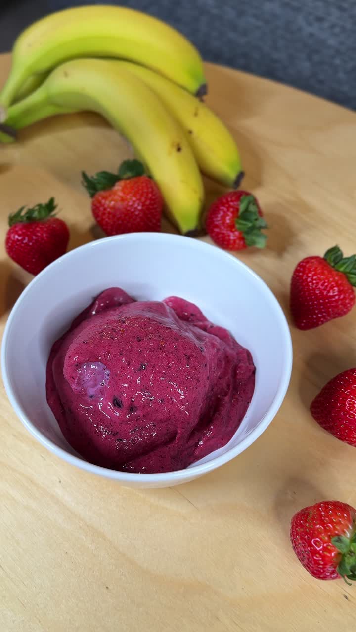 Una mano liscia uno spesso smoothie bowl all'açai in un piatto bianco, circondato da fragole e banane fresche: primo piano verticale e pulito, ideale per passaggi di ricette, colazioni salutari e promozioni benessere