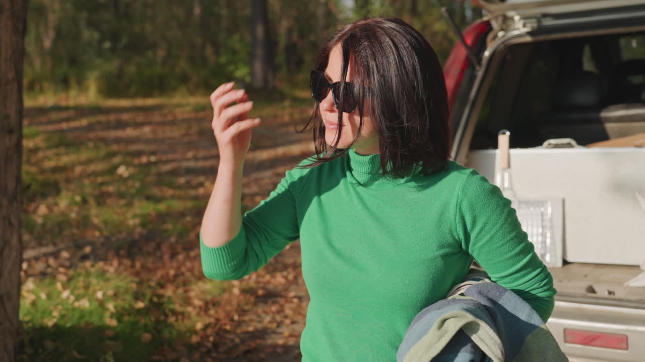 White woman unpacking car camping gear in autumn forest, wearing green sweater and jeans, removes rolled mat from suv trunk, carries supplies across leafstrewn ground, adjusts hair and ties sneakers