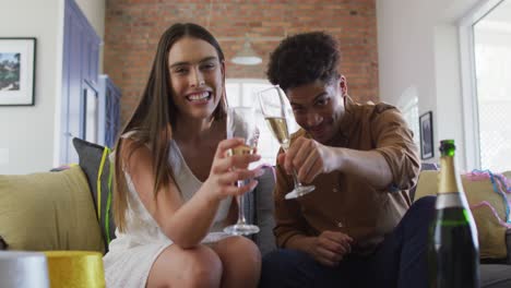 happy biracial couple drinking champagne at home making celebration laptop video call and laughing