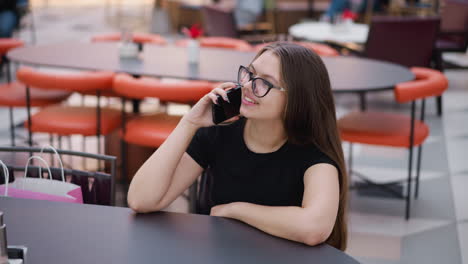 mujer joven con gafas en camiseta negra sentada en la mesa hablando por teléfono, sonriendo mientras disfruta de la conversación, con mesas y sillas cuidadosamente dispuestas en el fondo dentro de un moderno patio de comidas